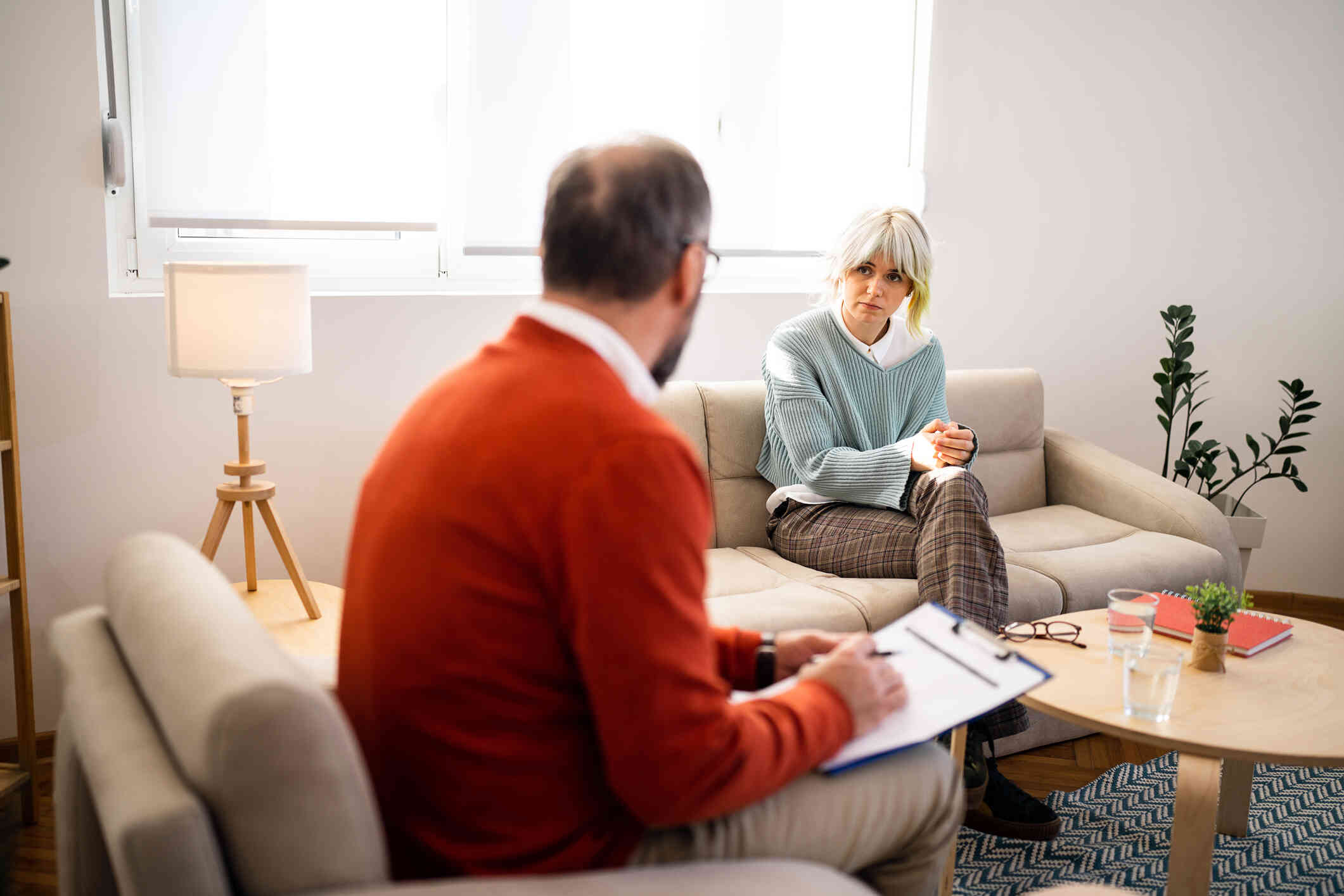 A woman in a blue sweater sits on a couch with her hands pressed together as she listens to the male therapist sitting across from her with a serious expression during a therapy session.